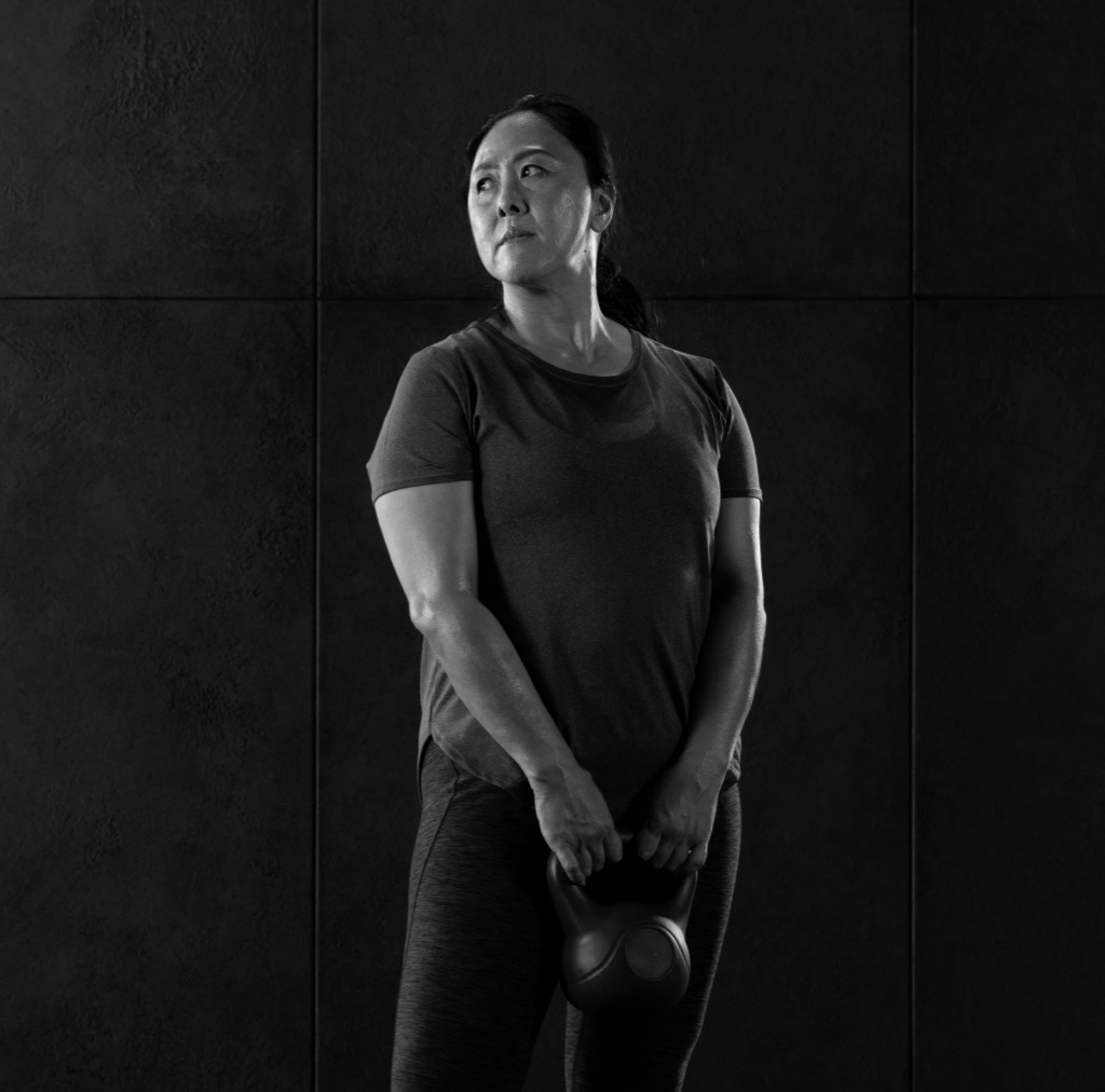 Imagem de uma mulher em trajes atléticos, segurando um kettlebell preto na altura da cintura. Ela está vestindo uma camiseta escura e leggings, com o cabelo preso em um rabo de cavalo. Sua expressão facial demonstra determinação ou foco, enquanto ela olha para cima e para a esquerda. O fundo é uma parede escura composta por grandes azulejos quadrados.