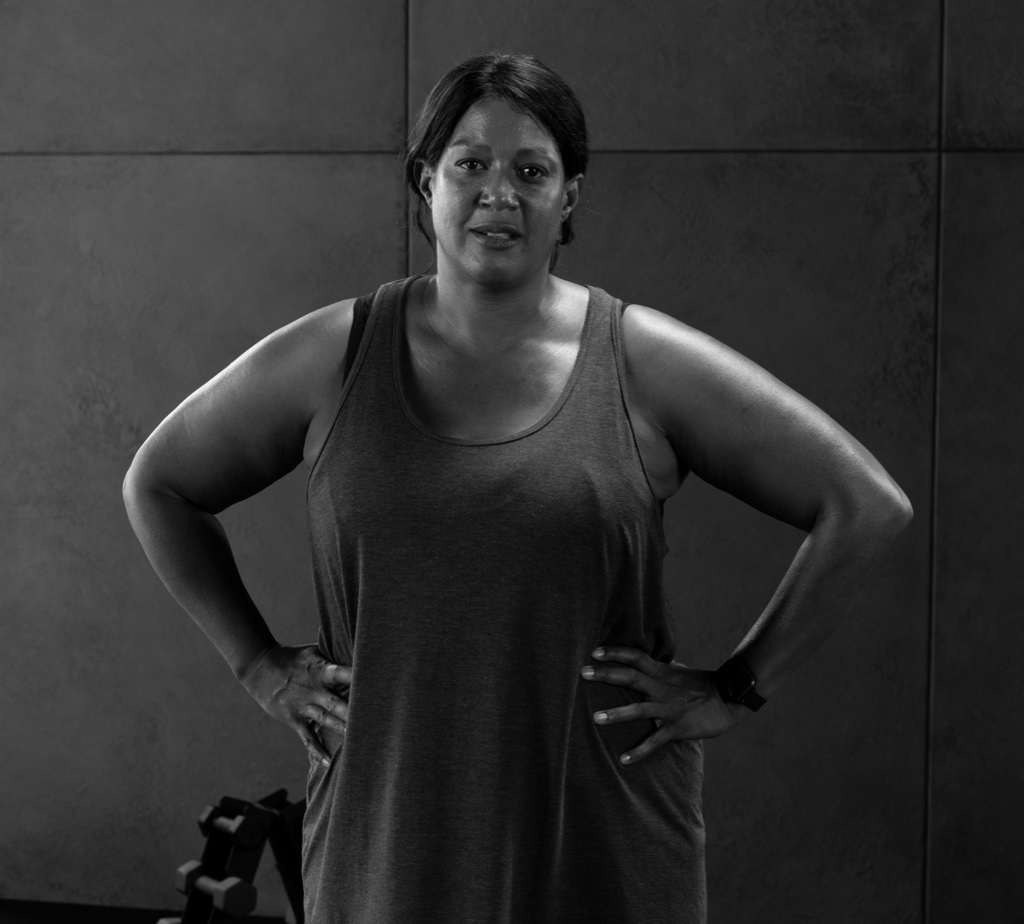 Fotografia em preto e branco de uma mulher em pé com as mãos nos quadris. Ela tem cabelo escuro preso para trás e está vestindo uma regata. Sua linguagem corporal sugere confiança e assertividade. A mulher parece estar em um ambiente de academia ou fitness, possivelmente após um treino. O fundo é uma parede lisa com um padrão de grade, composta por grandes azulejos ou painéis quadrados, que adicionam textura e profundidade à imagem. Há alguns objetos indistintos no fundo, que parecem ser equipamentos de exercício ou outros itens relacionados à academia, mas não são claramente visíveis devido ao baixo contraste e à falta de detalhes na imagem. A imagem transmite uma sensação de força e determinação, com a pose confiante da mulher e o ambiente de academia sugerindo um foco em fitness e bem-estar.